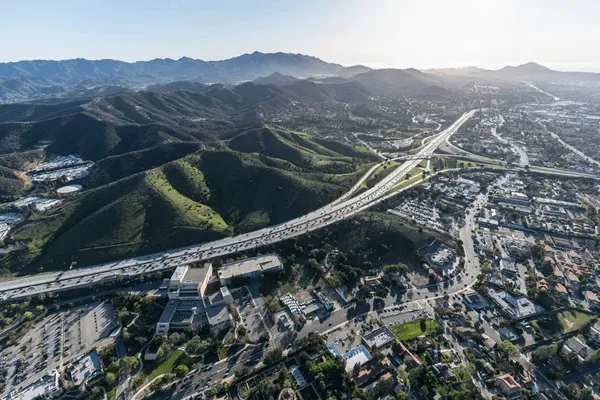 Aerial view of Ventura County, CA showing freeway, neighborhoods, and surrounding green hills under sunlight.