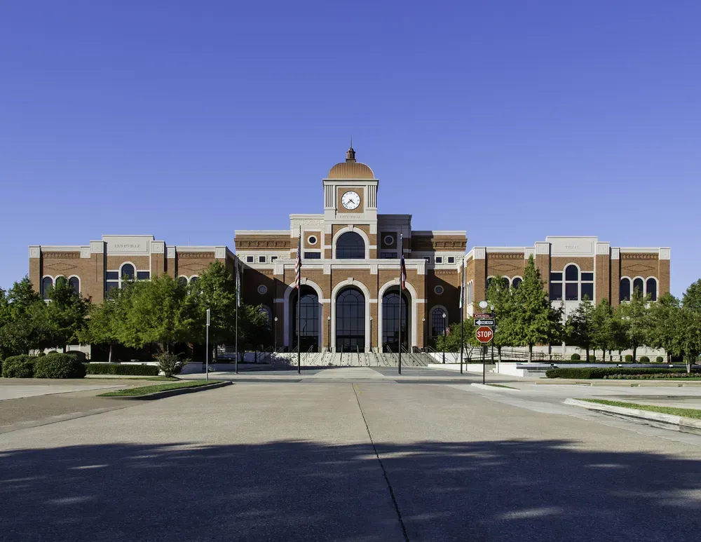 Lewisville City Hall in Lewisville, Texas, showcasing its red-brick architecture, clock tower, and surrounding trees on a clear sunny day.