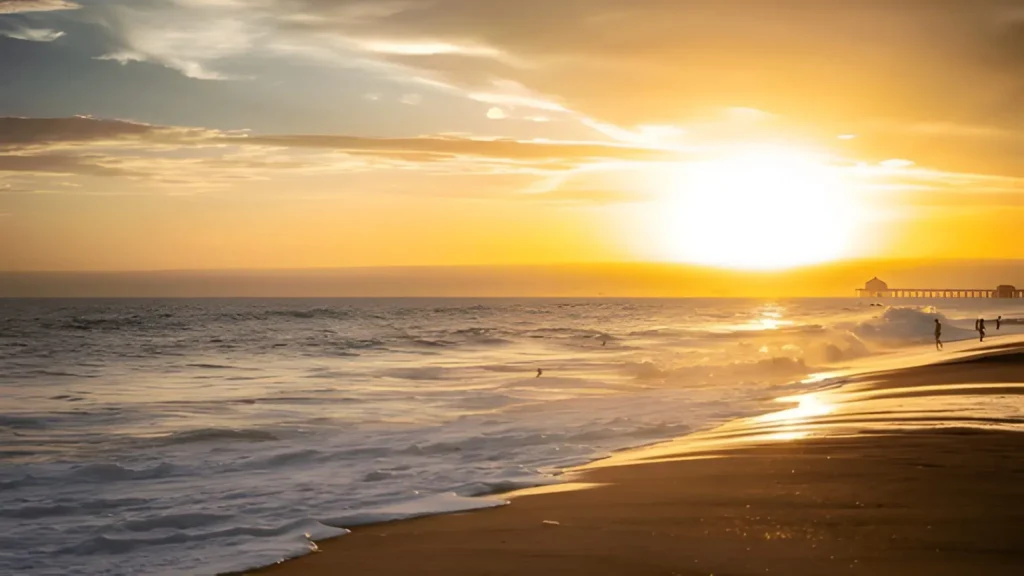 Sunset view over Huntington Beach, CA, with waves rolling onto the sandy shore and silhouettes of people walking along the beach near the pier.