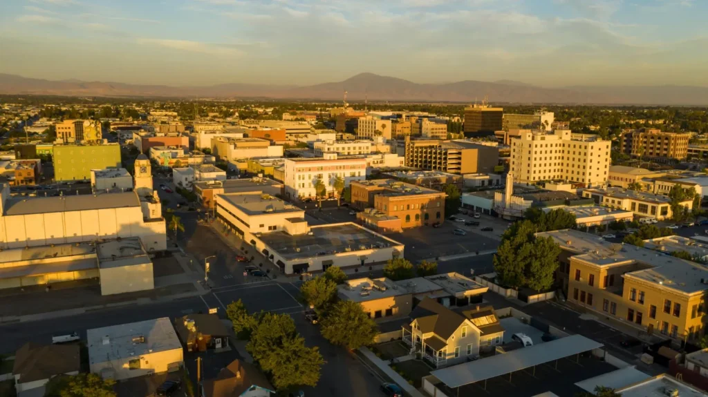 Aerial view of downtown Bakersfield in Kern County, California, at sunset showing city buildings, streets, and distant mountains in warm golden light.