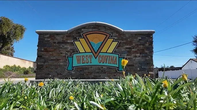 Welcome sign for West Covina, CA, displayed on a stone monument with yellow flowers in the foreground and a bright blue sky in the background.