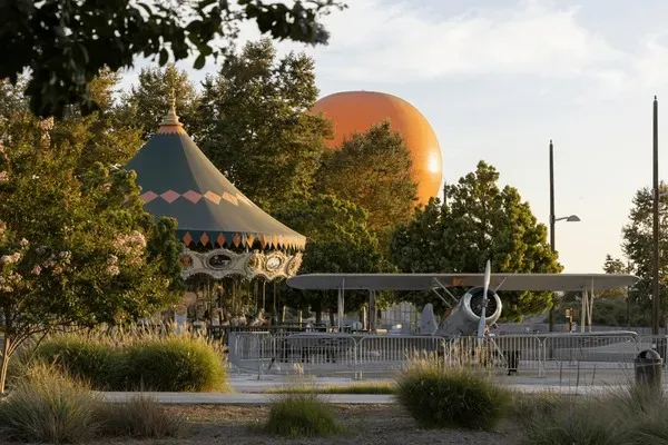 A small carousel and a model airplane are set among trees, with a large orange sphere visible in the background under a partly cloudy sky.