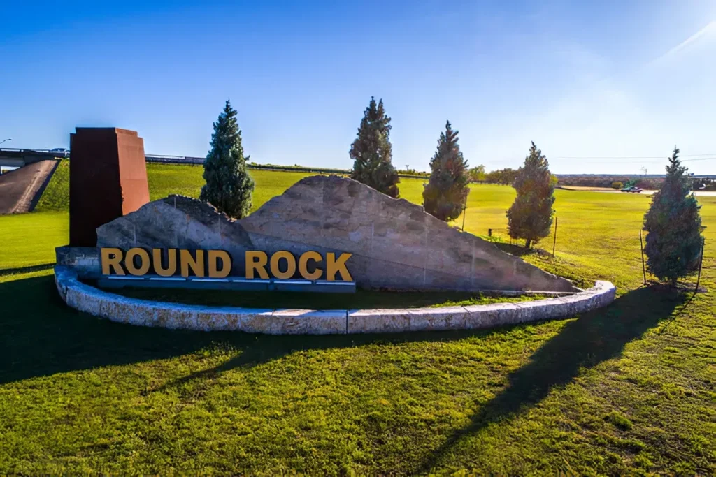 Round Rock TX city welcome sign near highway with stone sculpture and landscaped greenery under clear blue sky