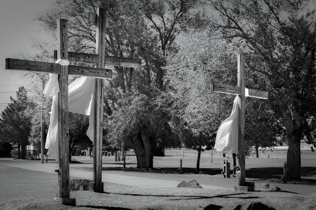 Three wooden crosses draped in white cloth at a public park in Clint, Texas, surrounded by trees and a quiet paved pathway.