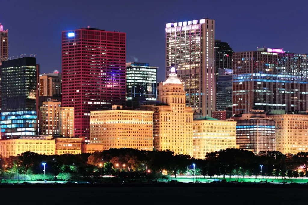 Night view of illuminated skyscrapers and office buildings in downtown Waco, Texas, with a park in the foreground and vibrant lights reflecting urban nightlife